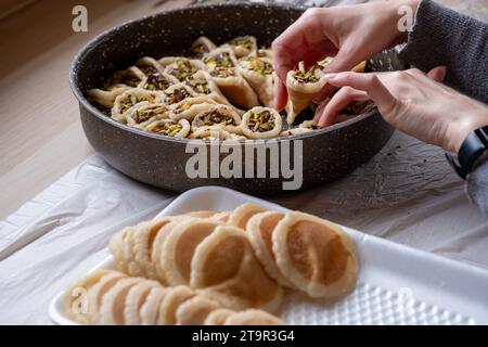 Hände halten Qatayef gefüllt mit Schokolade und mit Pistazien belegt auf einem Holztisch, mit einem Teller, der später im Ofen als Ramadan swe zubereitet wird Stockfoto