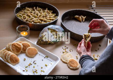 Hände halten Qatayef gefüllt mit Schokolade und mit Pistazien belegt auf einem Holztisch, mit einem Teller, der später im Ofen als Ramadan swe zubereitet wird Stockfoto