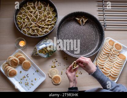 Hände halten Qatayef gefüllt mit Schokolade und mit Pistazien belegt auf einem Holztisch, mit einem Teller, der später im Ofen als Ramadan swe zubereitet wird Stockfoto