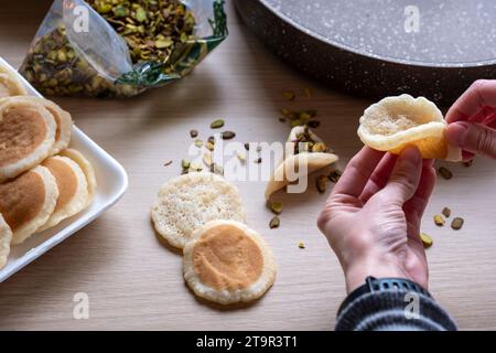 Hände halten Qatayef gefüllt mit Schokolade und mit Pistazien belegt auf einem Holztisch, mit einem Teller, der später im Ofen als Ramadan swe zubereitet wird Stockfoto