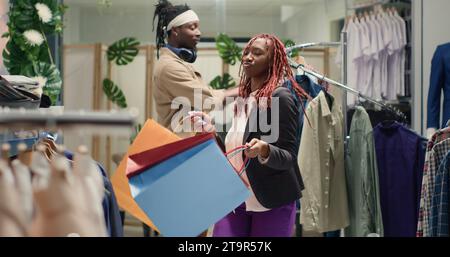 afroamerikanerin tanzt im Bekleidungsgeschäft mit Einkaufstaschen in der Hand und freut sich über Werbeaktionen. BIPOC-Kunde in einer Modeboutique, der Tanzbewegungen macht und sich über niedrige Preise freut Stockfoto