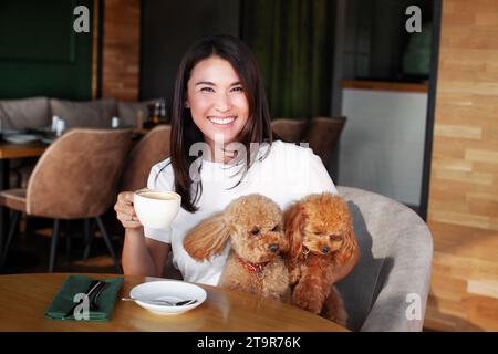 Porträt einer lächelnden Frau mit zwei kleinen Hundepudeln, die bei einer Tasse Kaffee am Tisch im Restaurant sitzen. Beziehungen zwischen Menschen und Haustieren. Schön Stockfoto