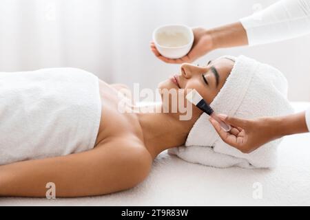 Entspannte junge indische Frau, die sich von Kosmetikerin Gesichtspflege macht Stockfoto