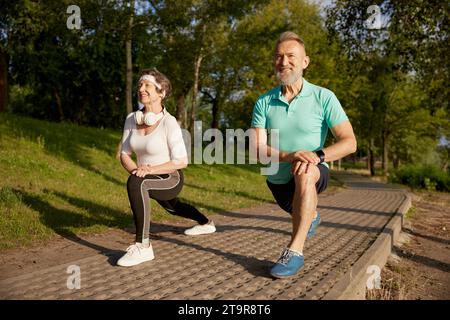 Lächelndes Seniorenpaar, das Ausfallschritte macht und sich vor dem Laufen die Beine aufwärmt Stockfoto
