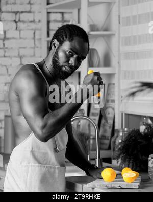 Schwarzer Mann, der zu Hause Frühstück oder Mittagessen in der Küche kocht. Afroamerikanischer Mann, der eine Schürze trägt und frische Zitrone zubereitet. Rohe Zutaten auf dem Tisch. Gesundheit Stockfoto
