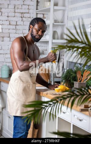 Schwarzer Mann, der zu Hause Frühstück oder Mittagessen in der Küche kocht. Afroamerikanischer Mann, der eine Schürze trägt und frische Zitrone zubereitet. Rohe Zutaten auf dem Tisch. Gesundheit Stockfoto
