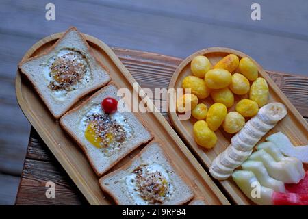 Ein Teller mit geröstetem Brot und Eiern, neben einem Teller mit gebackenen Kartoffeln, geschnittenen Bananen, Melonen und Wassermelonen. Stockfoto