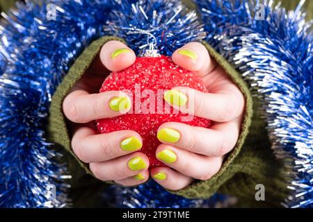 Die Hände der Frau halten einen rot glänzenden Weihnachtsball. Modische grüne Maniküre, Lametta. Weihnachtsfeier und Neujahrsfest Stockfoto