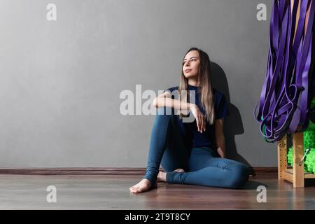 Eine junge Frau ruht sich nach einem Fitnesstraining aus, sitzt mit dem Rücken an der Wand in Sportbekleidung, ein durchgehendes Porträt mit einer Kopie des Raumes Stockfoto