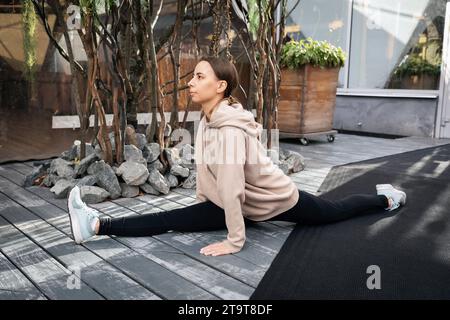 Eine junge, sportliche, braunhaarige Frau spielt Sport auf der Straße, führt eine Übung durch, um die Muskeln ihrer Beine zu dehnen, sitzt in einer Längsspalte, Stockfoto