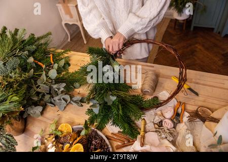 Draufsicht Komposition weiblicher Hände, die Weihnachtskranz aus natürlichen Zweigen von Fichte, Kiefer, Eukalyptus herstellen. Das Mädchen wickelt grüne Zweige auf die Kraft-Basis Stockfoto