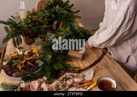 Draufsicht Komposition weiblicher Hände, die Weihnachtskranz aus natürlichen Zweigen von Fichte, Kiefer, Eukalyptus herstellen. Das Mädchen wickelt grüne Zweige auf die Kraft-Basis Stockfoto