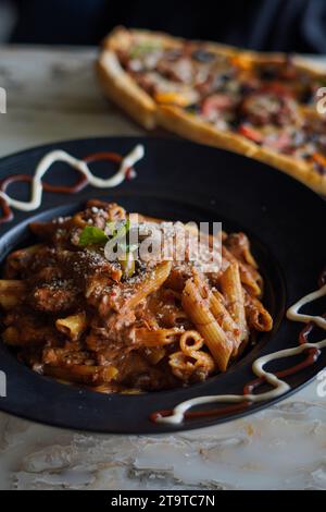 Penne Pasta mit Sauce, Oliven und frisch geriebenem Parmesankäse, auf einer schwarzen Platte, mit Sauce an der Seite. Und eine frische Pizza im Hintergrund. Stockfoto