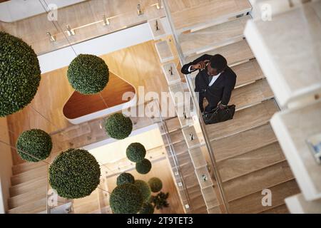 Top-down-Aufnahme eines beschäftigten afroamerikanischen Geschäftsmannes mit Ledertasche, der telefonisch anruft, der auf einer Treppe in einem modernen Bürogebäude steht, Kopierraum Stockfoto