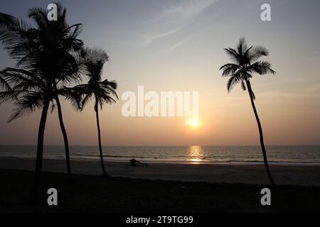 Eine Silhouette aus Kokospalmen und ein Fischerboot am Strand bei Sonnenuntergang am Colva Beach in Goa, Indien Stockfoto
