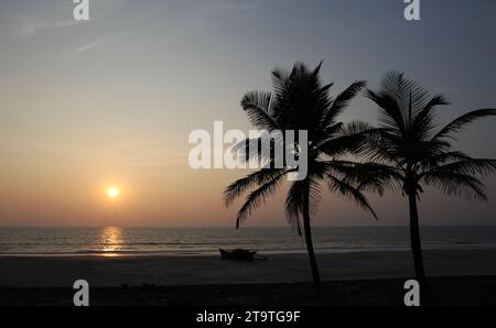 Eine Silhouette aus Kokospalmen und ein Fischerboot am Strand, während die Sonne am Colva Beach in Goa, Indien, untergeht Stockfoto