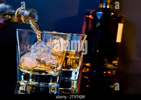 Der Barkeeper gießt Whiskey in zwei Gläser mit Eiswürfeln auf dem Tisch mit hellblauem Licht und Reflexion, Zeit der Entspannung mit Whisky Stockfoto