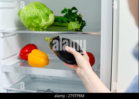 Eine Frau stellt eine Aubergine in den Kühlschrank. Frisches Gemüse liegt auf dem Regal im Kühlschrank. Stockfoto