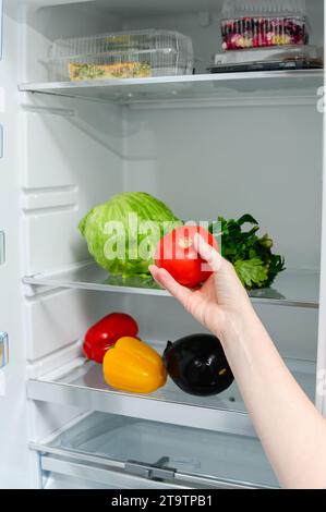 Frisches Gemüse liegt auf dem Regal im Kühlschrank. Die Hand einer Frau hält eine Tomate vor dem Hintergrund eines Kühlschranks. Stockfoto