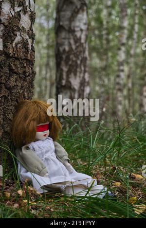 Gruselige Puppe mit roter Blindfalte, die im Wald sitzt. Mystery, Thriller, Horror, Vibes. Stockfoto