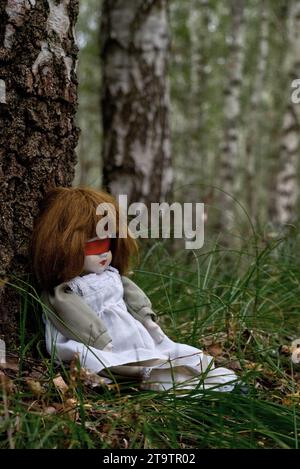 Gruselige Puppe mit roter Blindfalte, die im Wald sitzt. Mystery, Thriller, Horror, Vibes. Stockfoto