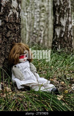 Gruselige Puppe mit roter Blindfalte, die im Wald sitzt. Mystery, Thriller, Horror, Vibes. Stockfoto