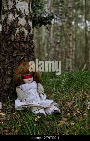 Gruselige Puppe mit roter Blindfalte, die im Wald sitzt. Mystery, Thriller, Horror, Vibes. Stockfoto