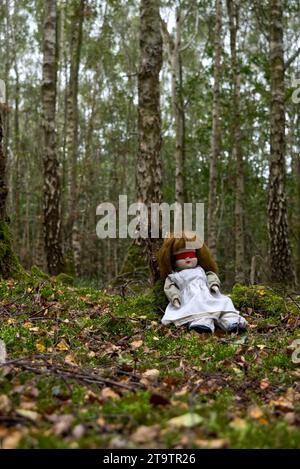 Gruselige Puppe mit roter Blindfalte, die im Wald sitzt. Mystery, Thriller, Horror, Vibes. Stockfoto