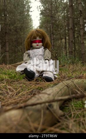 Gruselige Puppe mit roter Blindfalte, die im Wald sitzt. Mystery, Thriller, Horror, Vibes. Stockfoto