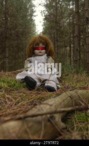 Gruselige Puppe mit roter Blindfalte, die im Wald sitzt. Mystery, Thriller, Horror, Vibes. Stockfoto