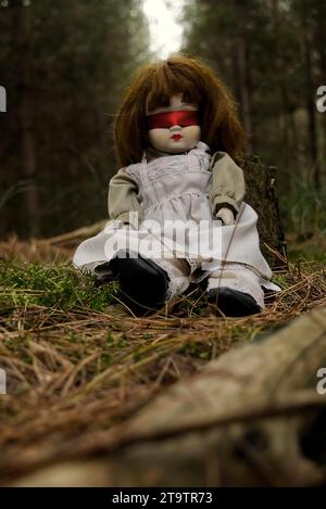 Gruselige Puppe mit roter Blindfalte, die im Wald sitzt. Mystery, Thriller, Horror, Vibes. Stockfoto