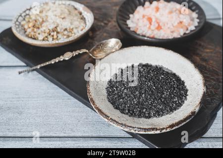 Verschiedene Salzsorten. Drei verschiedene Salzsorten in handgefertigten Tellern auf einer Tonplatte. Weißes Kräutersalz, rosa Himalaya und schwarzer Maldo Stockfoto