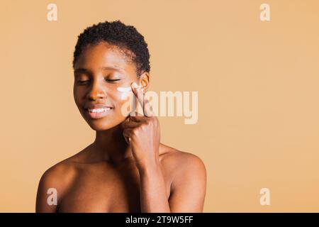 Junge, schwarz lächelnde Frau mit geschlossenen Augen, die Feuchtigkeitscreme auf ihr Gesicht aufträgt. Junge Frau, die ihre Gesichtshaut mit einem Schönheitsprodukt behandelt. Stockfoto