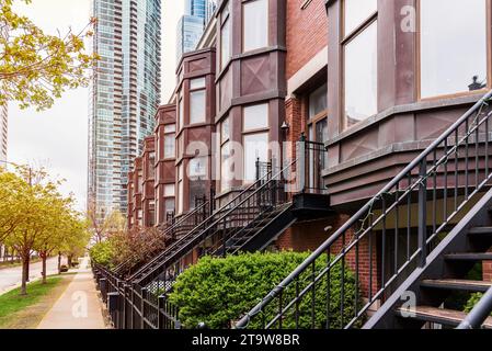 Reihenhäuser mit einer Metalltreppe, die zur Tür führt, entlang eines von Bäumen gesäumten Gehwegs. Im Hintergrund befinden sich Hochhäuser. Stockfoto