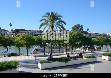 Sevilla, Spanien - 25. November 2023: Menschen auf einer Bank auf einem Fußweg entlang des Flusses Guadalquivir mit Blick auf das Triana-Viertel in Sevilla, Spanien. Stockfoto
