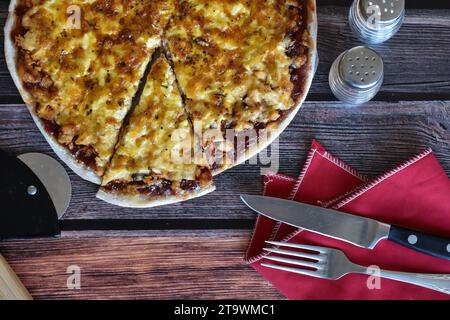 Frisch gebackene hausgemachte Pizza auf Holztisch. Hähnchenpizza mit Käse, Pizzascheibe. Pizzaschneider, rote Serviette, Messer und Gabel, Salz- und Pfefferstreuer. Stockfoto