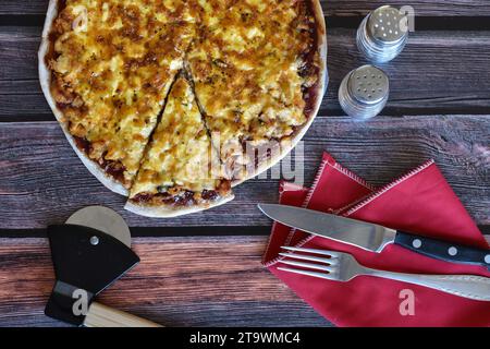 Frisch gebackene hausgemachte Pizza auf Holztisch. Hähnchenpizza mit Käse, Pizzascheibe. Pizzaschneider, rote Serviette, Messer und Gabel, Salz- und Pfefferstreuer. Stockfoto