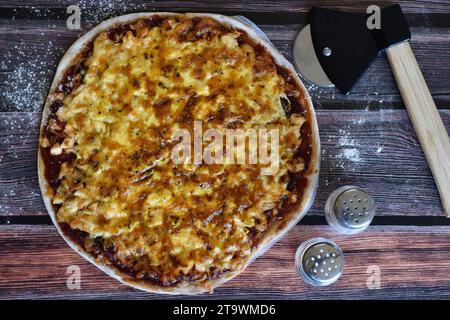 Frisch gebackene, hausgemachte Pizza, serviert auf einem bemehlten Holztisch. Leckere Hühnerpizza mit Käse. Pizzaschneider, Salz- und Pfefferstreuer. Draufsicht. Stockfoto