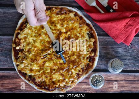 Die Hand des Mannes schneidet hausgemachte Pizza auf Scheiben mit einem Spezialmesser. Hühnchenkäse-Pizza auf Holztisch. Rote Serviette, Messer und Gabel, Salz und Pfeffer. Stockfoto