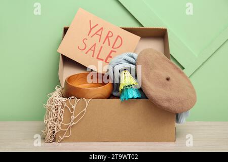 Schachtel mit unerwünschtem Zeug und Pappschild mit Text YARD SALE auf grünem Hintergrund Stockfoto
