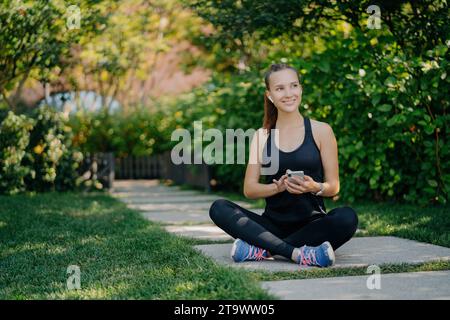 Glückliche junge Frau, die im Kreuz auf einem Weg in einem Park sitzt, ein Smartphone hält und Sportkleidung trägt Stockfoto