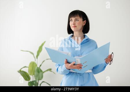 Professionelle Geschäftsfrau im formellen blauen Hemd prüft Papierdokumente in der Mappe steht auf weißem Wandhintergrund mit großer grüner Pflanze. Rechtsanwalt, Rechtsabteilung Stockfoto