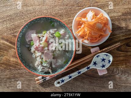 Heißer gekochter Reis oder Reisbrei mit Schweinerippchen Suppe und Schinken serviert mit eingelegten Karotten und Rettich auf hölzernem Schneidebrett. Gesundes Frühstück Stockfoto