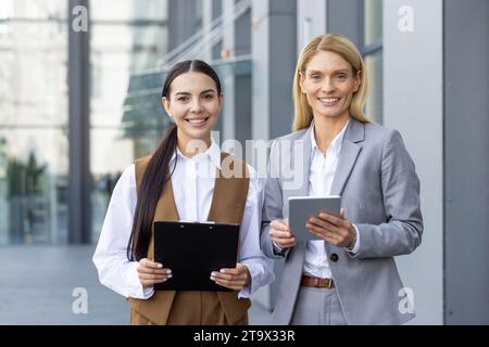 Porträt von zwei Geschäftsfrauen, die vor dem Bürozentrum in Geschäftskleidung stehen, ein Tablet und einen Ordner mit Dokumenten in der Hand halten und in die Kamera schauen und lächeln. Stockfoto
