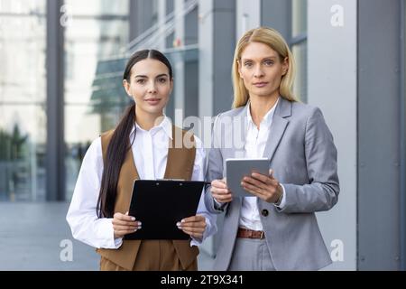 Zwei Geschäftsfrauen stehen auf der Straße in der Nähe eines Bürogebäudes, schauen ernsthaft in die Kamera, halten einen Ordner mit Dokumenten und ein Tablet in der Hand. Nahaufnahme Porträt. Stockfoto