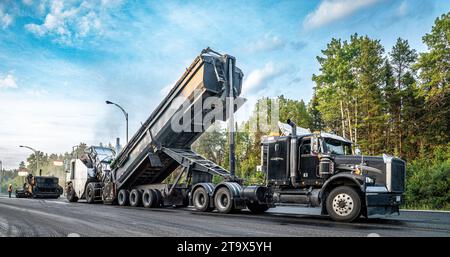 Sattelzugwagen entladen Asphalt in ein Transportfahrzeug während der Pflasterarbeiten auf dem Trans-Canada Highway, spät am Tag Stockfoto