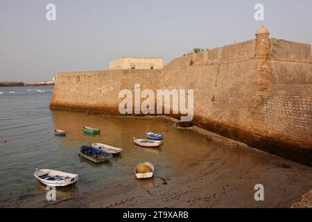 Fischerboote, die an den Mauern des Castillo de Santa Catalina am Strand in Cadiz, Spanien, ankern. Die pentagon-förmige Burg wurde gebaut, nachdem die Engländer 1596 die Stadt geplündert hatten, um die Bucht von Cadiz zu bewachen. Stockfoto