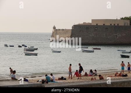 Strandgäste entspannen sich in der Nähe des kleinen Fischerboothafens an den Mauern des Castillo de Santa Catalina in Cadiz, Spanien. Der öffentliche Strand liegt zwischen zwei alten Burgen an der Bucht. Stockfoto