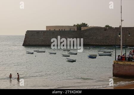 Strandgäste waten ins Wasser am kleinen Fischerboothafen entlang der Mauern des Castillo de Santa Catalina in Cadiz, Spanien. Der öffentliche Strand liegt zwischen zwei alten Burgen an der Bucht. Stockfoto