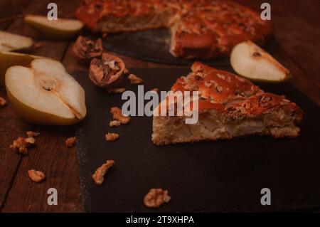 Birnenkuchen auf einem schwarzen Servierbrett auf einem Holztisch, geschnittene Birnen und Walnüsse liegen chaotisch Stockfoto